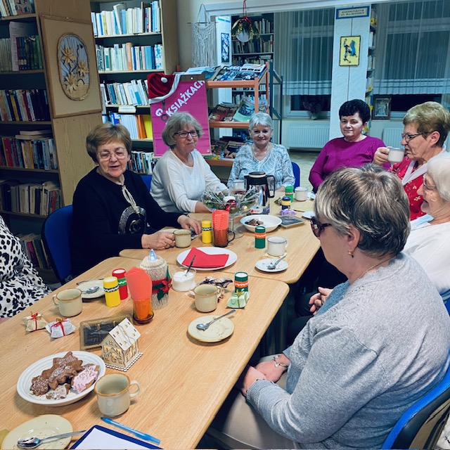 Uczestniczki spotkania siedzą przy stole i rozmawiają o przeczytanej książce. W tle plakat promocyjny Dyskusyjnych Klub&oacute;w Książki.