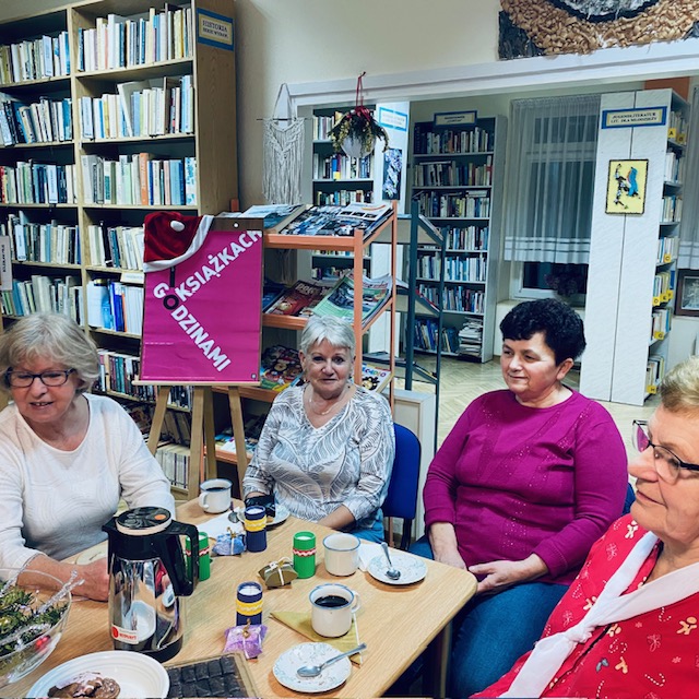 Uczestniczki spotkania siedzą przy stole i rozmawiają o przeczytanej książce. W tle plakat promocyjny Dyskusyjnych Klub&oacute;w Książki.