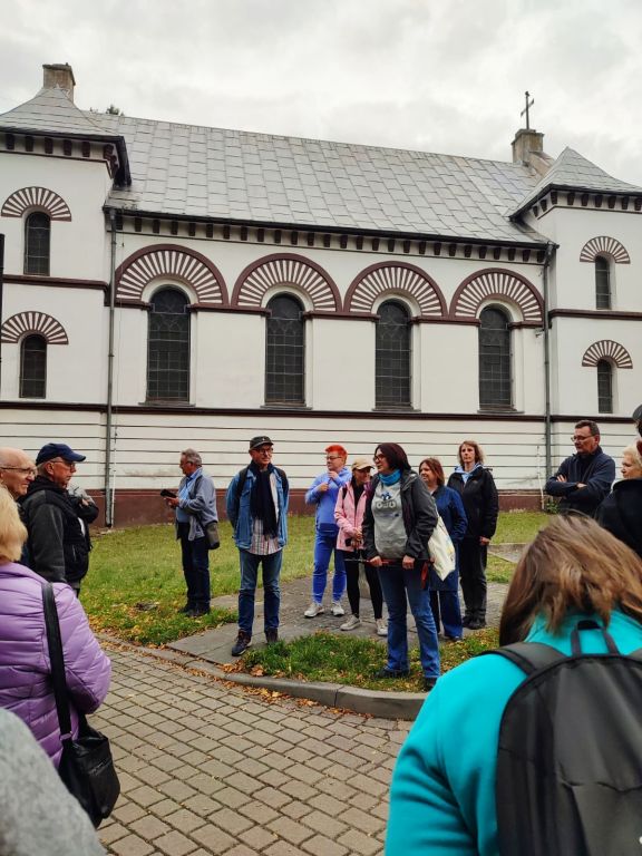 Grupa uczestników spaceru historycznego stoi na placu przed zabytkowym kościołem w Ozimku