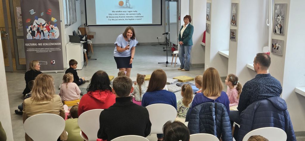 Młoda bibliotekarka nachyla się do dzieci siedzących na podłodze, które próbują rozwiązać zagadkę wyświetloną na ekranie multimedialnym.kę
