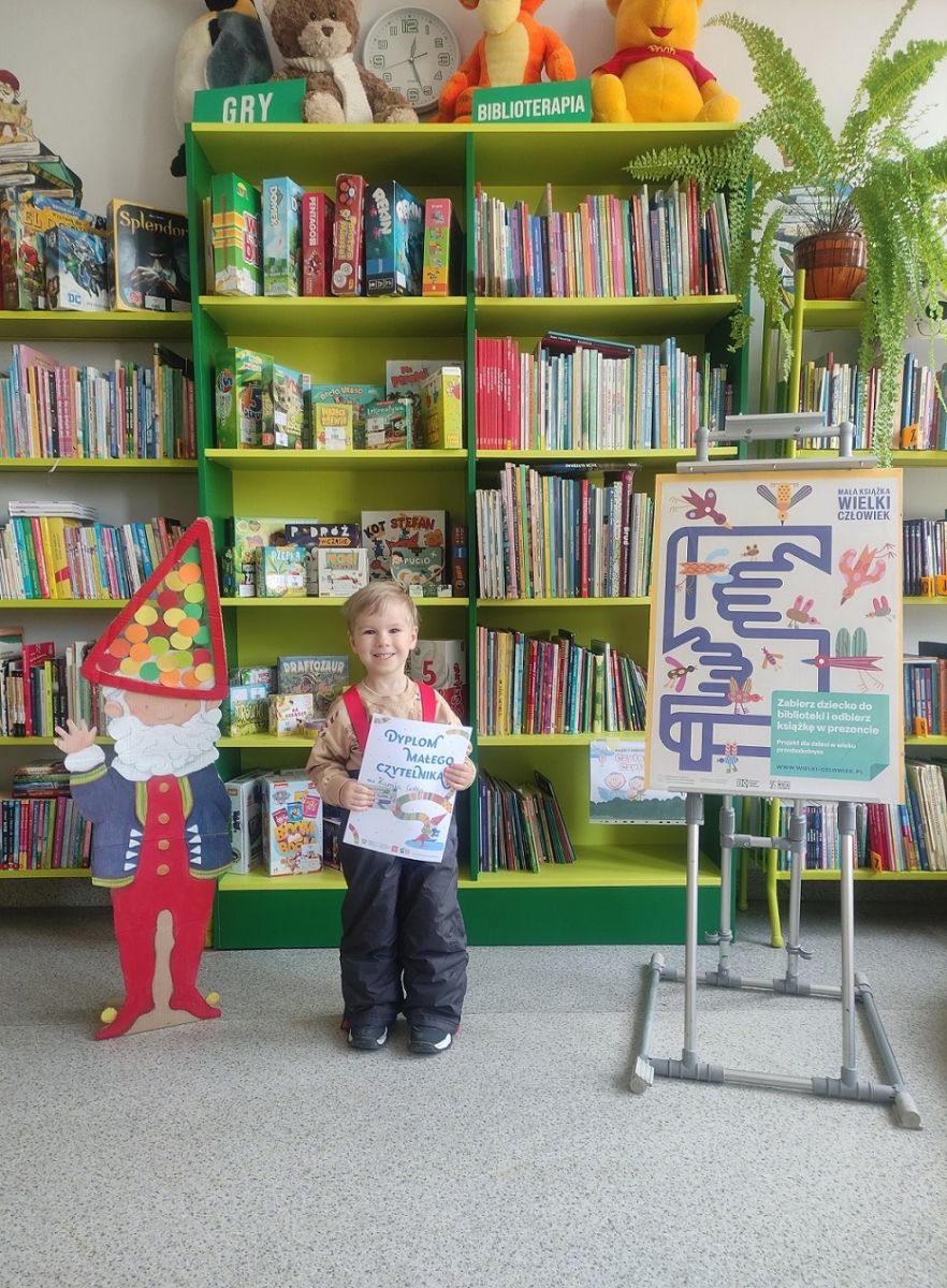 Chłopczyk stoi na tle p&oacute;łek z książkami i trzyma w rękach dyplom Małego Czytelnika.