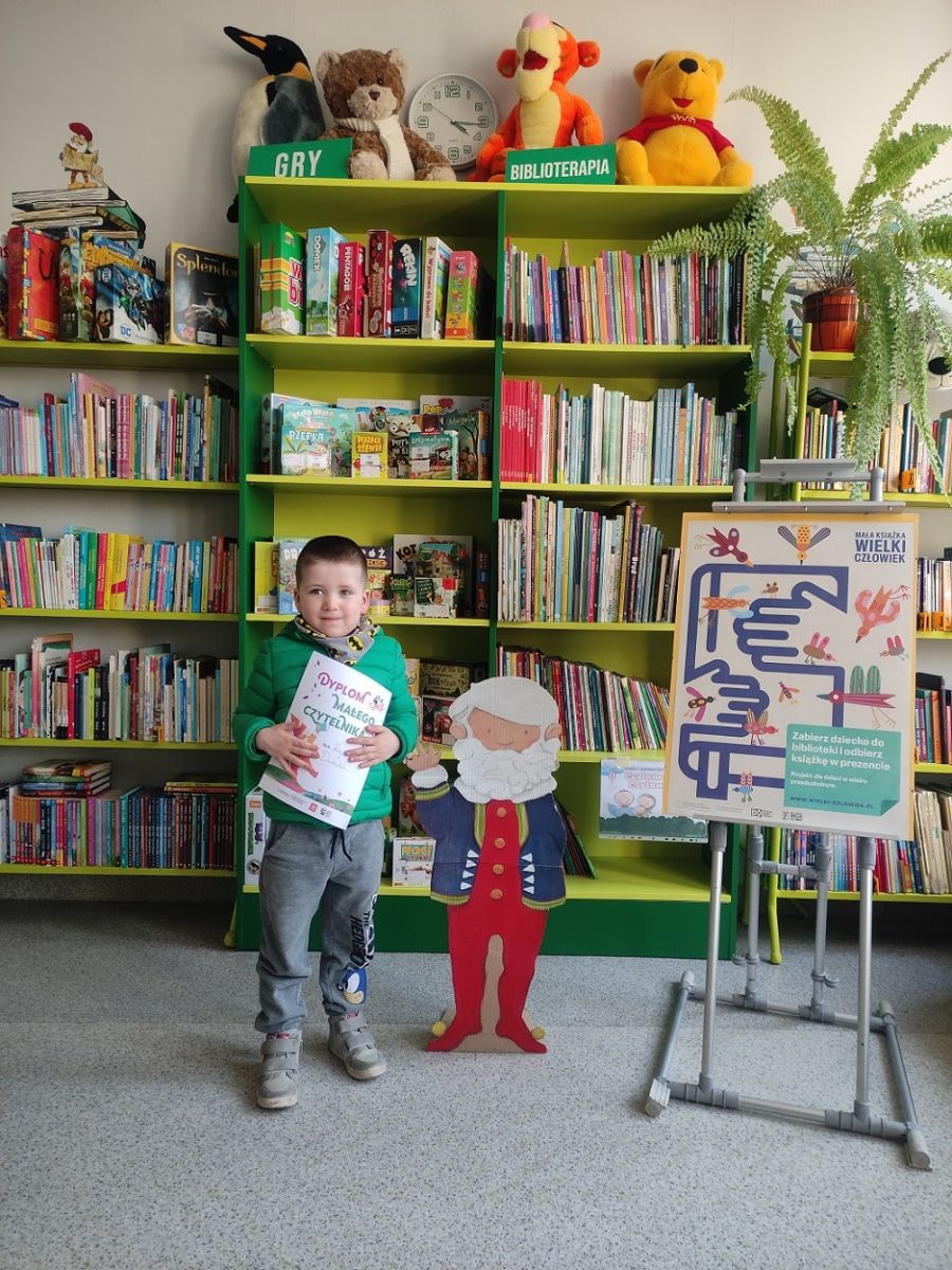 Chłopczyk stoi z dyplomem na tle regał&oacute;w z książkami. Obok stoi plakat promujący kampanię czytelniczą.