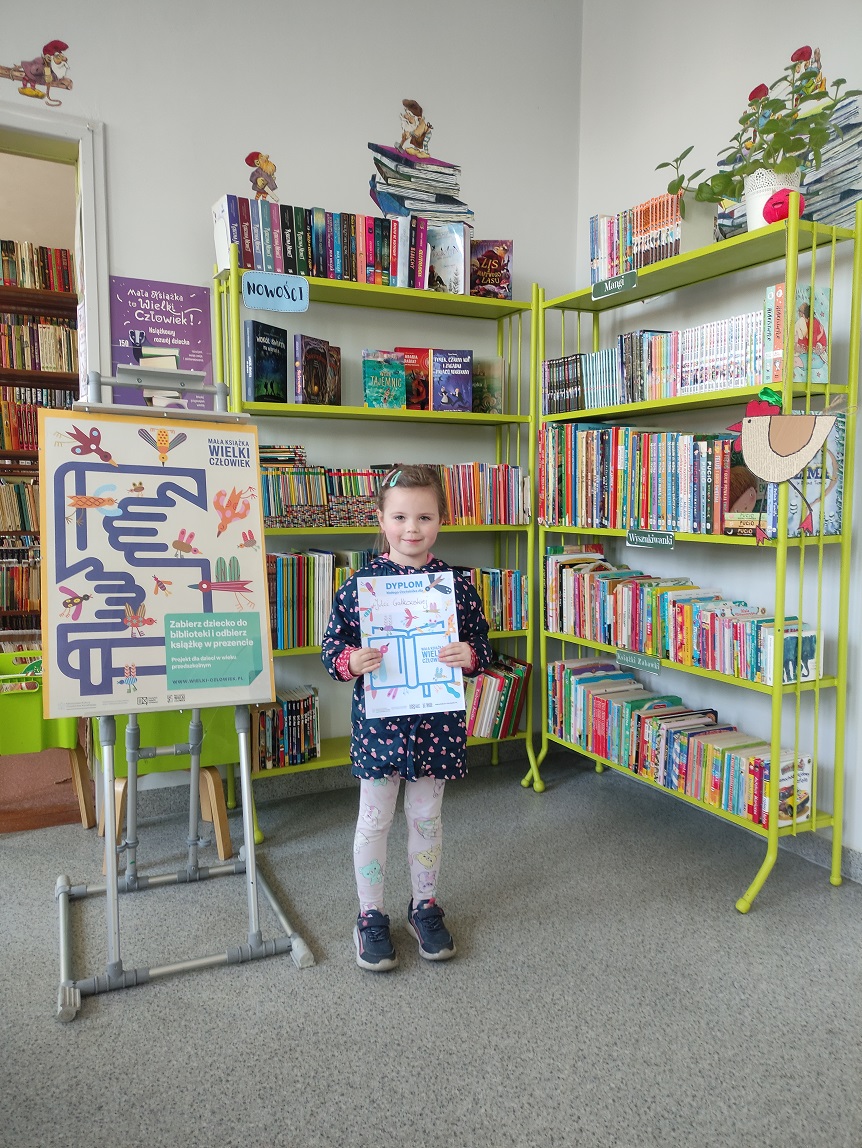 Dzieczynka stoi z dyplomem na tle regał&oacute;w z ksiązkami. Obok stoi plakat promujący kampanię czytelniczą.