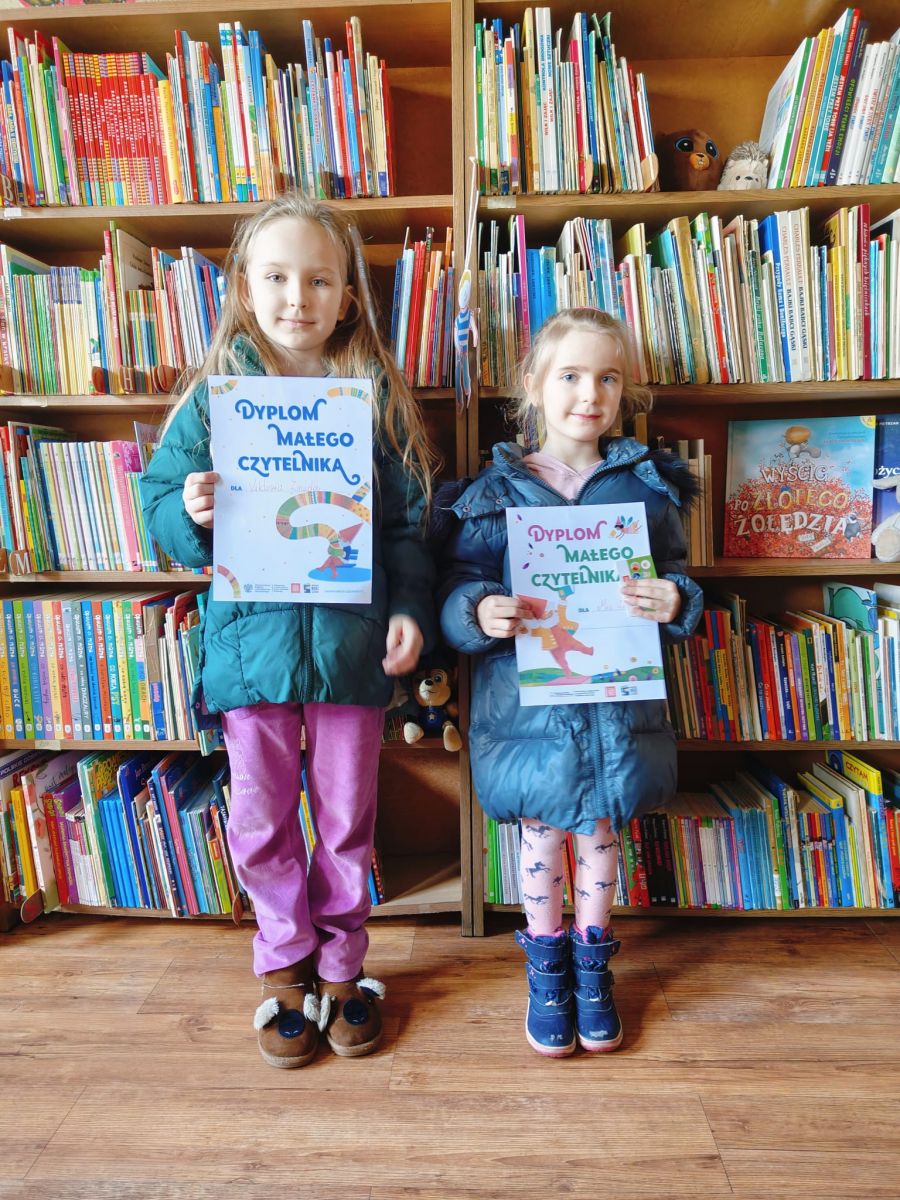 Dwie dziewczynki pozują do zdjęcia trzymając dyplom z projektu "Mała książka- wielki człowiek" na tle regał&oacute;w z książkami. 
