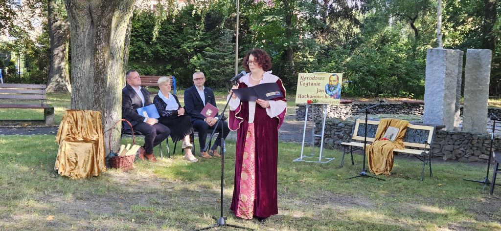 Kobieta ubrana w renesansową suknię wita gości na Narodowym Czytaniu. W tle na skwerku osoby przygotowujące się do czytania