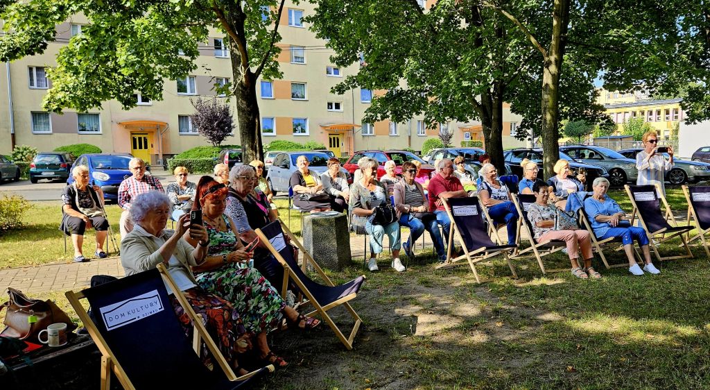 Grupa kilkudziesięciu osób siedzi na leżakach i krzesłach na skwerku i słucha utworów Jana Kochanowskiego
