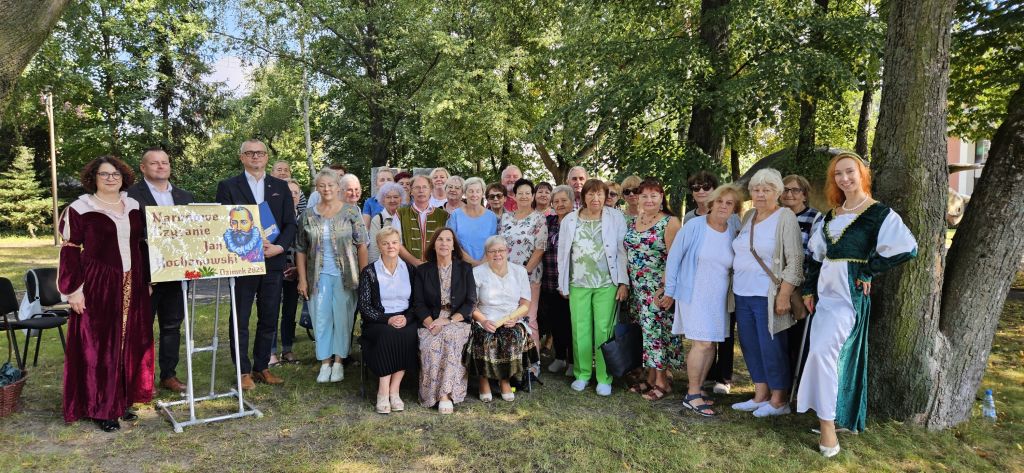 Uczestnicy spotkania i lektorzy stoją na skwerku przy tablicy z napisem Narodowe Czytanie Jan Kochanowski Ozimek 2025