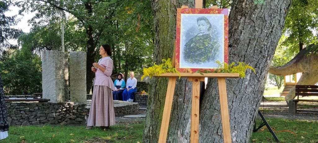 Kobieta ubrana w str&oacute;j z XIX wieku stoi w parku, widać zbliżenie zdjęcia Elizy Orzeszkowej