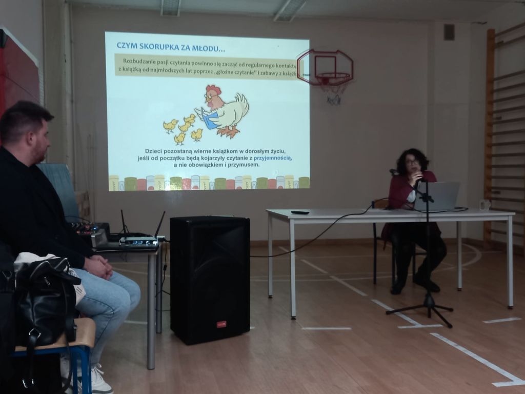 Kierowniczka biblioteki siedzi przy stole w sali gimnastycznej szkoły i wygłasza  prelekcje na temat czytania. W tle slajd prezentacji multimedialnej.