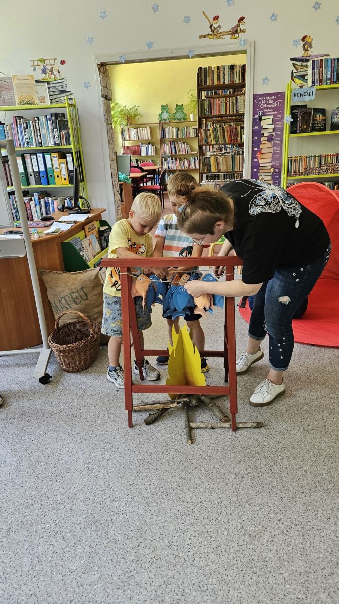 Dw&oacute;ch chłopc&oacute;w pomaga bibliotekarce zawiesić złowione ryby nad ogniskiem. W tle regały z książkami.