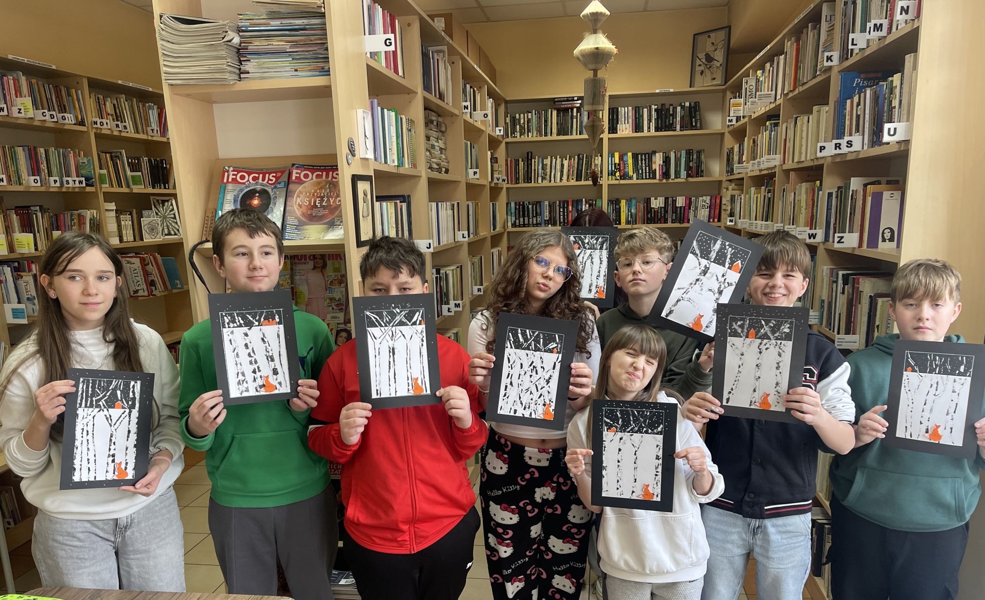Cała klasowa grupa, stojąc na tle regał&oacute;w i  eksponuje przed sobą swoje obrazki. 