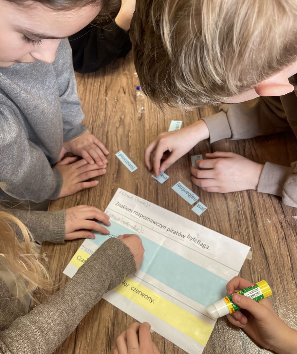 Dzieci pochylone nad kartką, na kt&oacute;rą doklejają rozsypanki wyrazowe, budując poprawne zdania.
