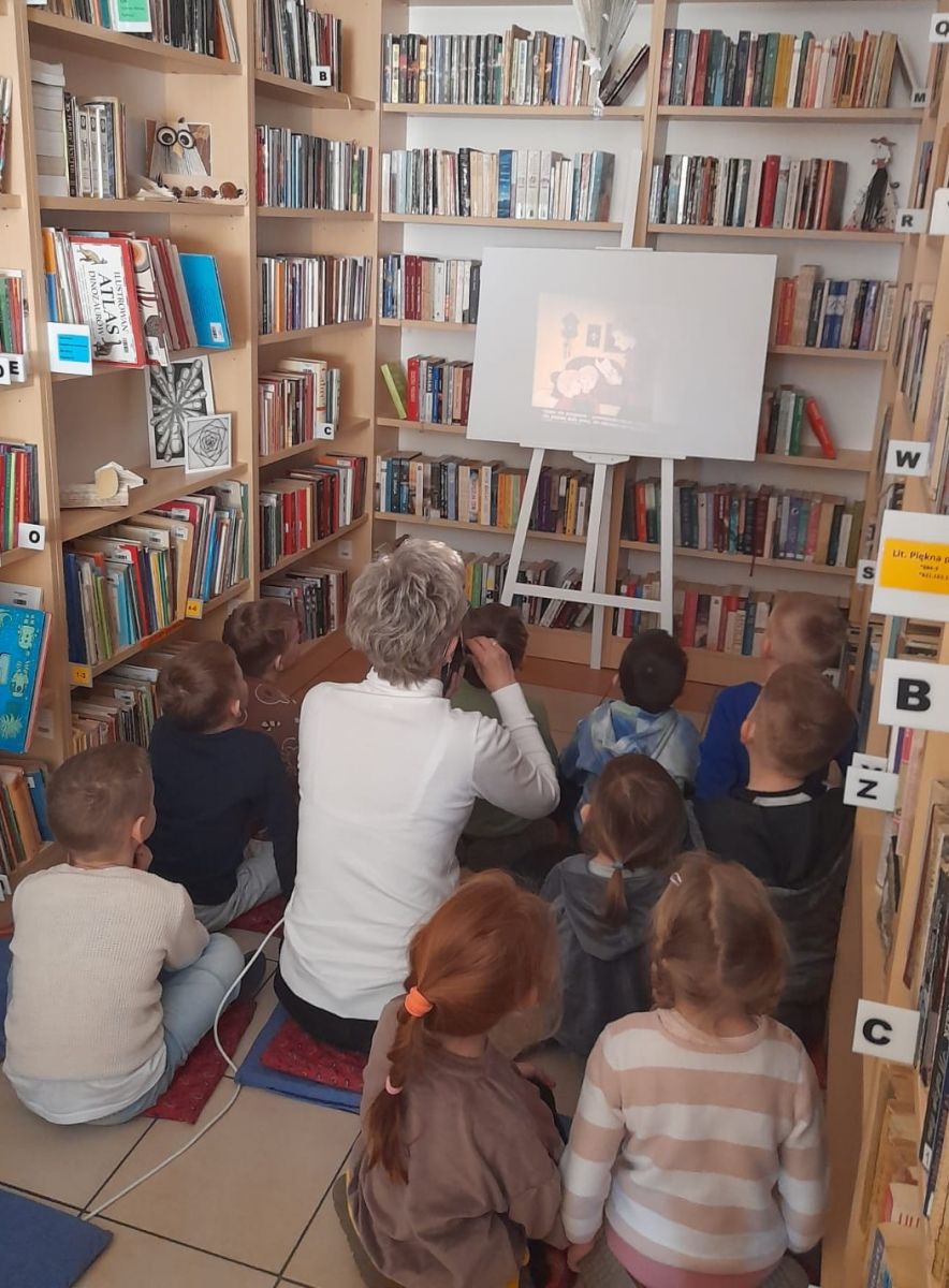 Przedszkolaki siedząc na podłodze wokoło bibliotekarki oglądają na ekranie bajkę, wyświetlaną za pomocą rzutnika..