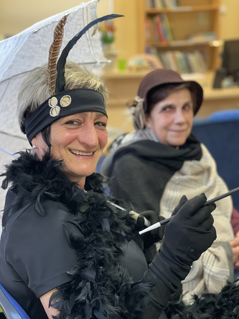 Zdjęcie portretowe dw&oacute;ch pań. Pierwsza, siedząc pod białym, koronkowym parasolem ma na głowie czarną opaskę, ozdobioną broszką i pi&oacute;rami, a wokoło szyi owinięty ma  pierzasty szal boa. W dłoni  trzyma długą cygaretkę. W tle, jej sąsiadka w subtelnym kapelusiku zerka z uśmiechem w obiektyw.