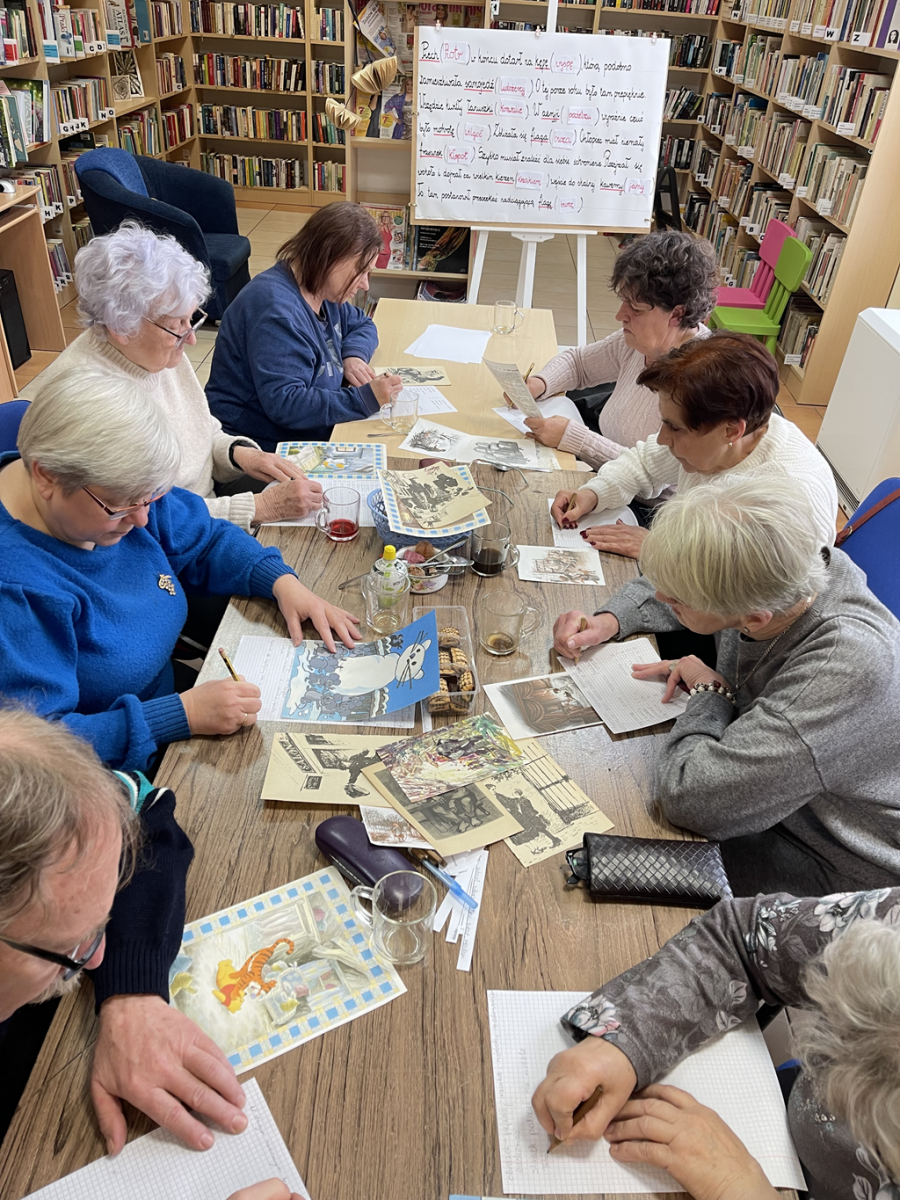 Ośmiu uczestnik&oacute;w spotkania, siedząc przy stołach pochyleni nad kartkami, redagują teksty literackie do wybranych wcześniej ilustracji.