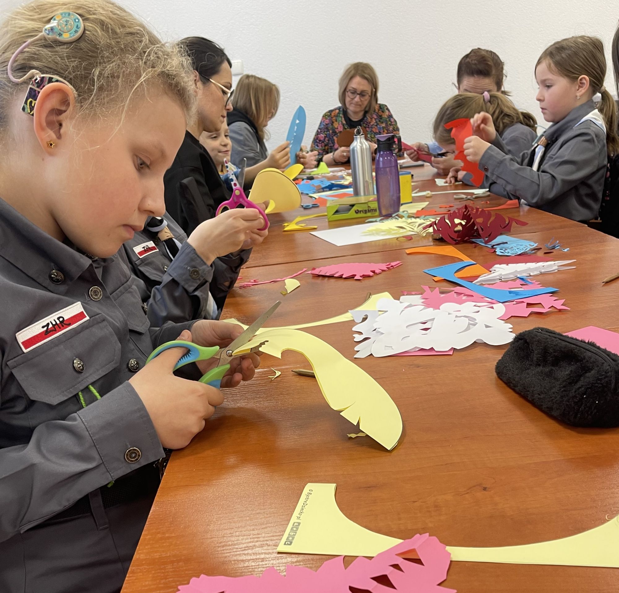 Przy podłużnym stole, na blacie kt&oacute;rego jest mn&oacute;stwo kolorowych kartek, siedzą dzieci i ich mamy. Wszyscy w skupieniu zajęci są pracą nad własną wycinanką.