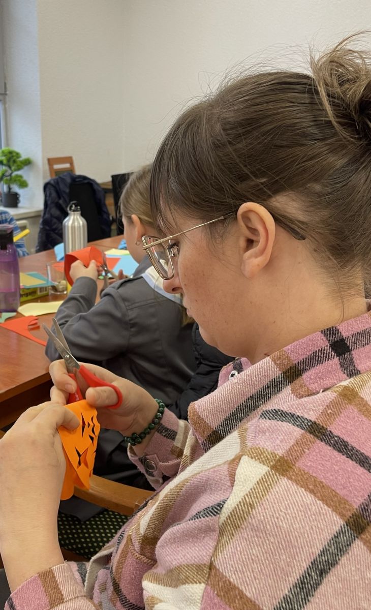 Zbliżenie na profil kobiety, pracującej nad wycinanką. W tle siedzi dziewczynka r&oacute;wnież pochylona nad swoją pracą.