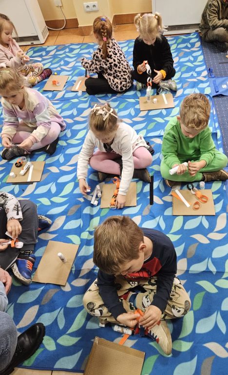 Grupa przedszkolak&oacute;w, siedząca na dywanie, skleja kolorowe paski  w papierowe łańcuchy, kt&oacute;re będą mackami ośmiornicy.
