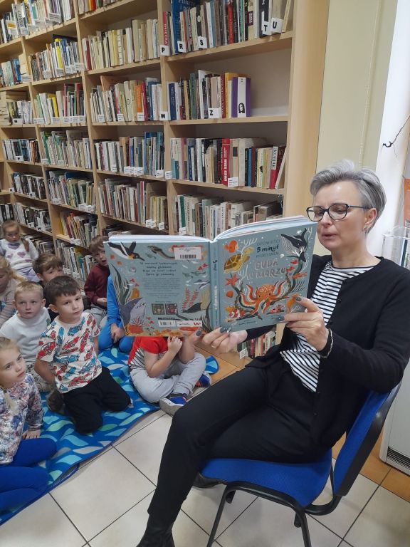 Bibliotekarka, siedząca przed dziećmi z uniesionej wysoko książki, czyta głośno tekst opowiadania. 