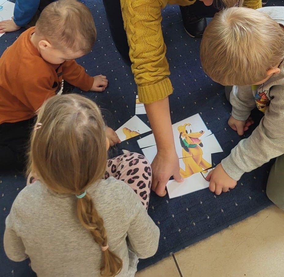 Tr&oacute;jka dzieci siedząca na dywanie układa puzzle z wizerunkiem psa Pluto.