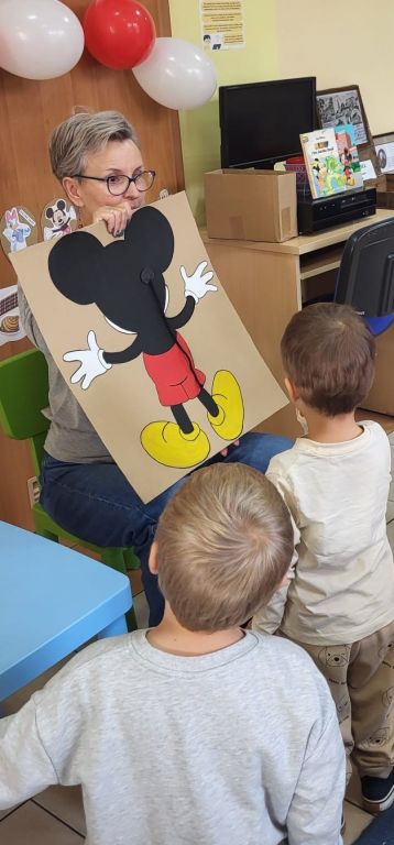 Bibliotekarka, mając przed sobą dw&oacute;jkę dzieci, trzyma opartą o swoje kolana planszę z wizerunkiem Myszki Miki i doklejonym nieporadnie ogonkiem.  