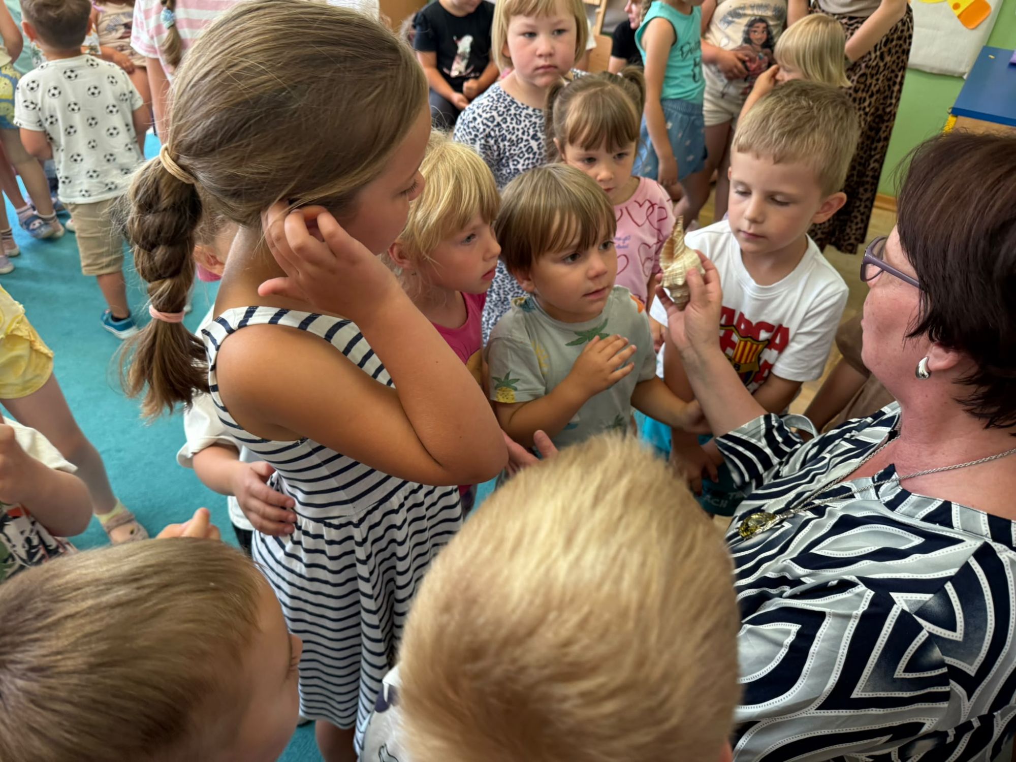 Pani podaje muszle zgroamdzonym wok&oacute;ł niej dzieciom, kt&oacute;re przykładają ją do ucha i słuchają szumu morza.