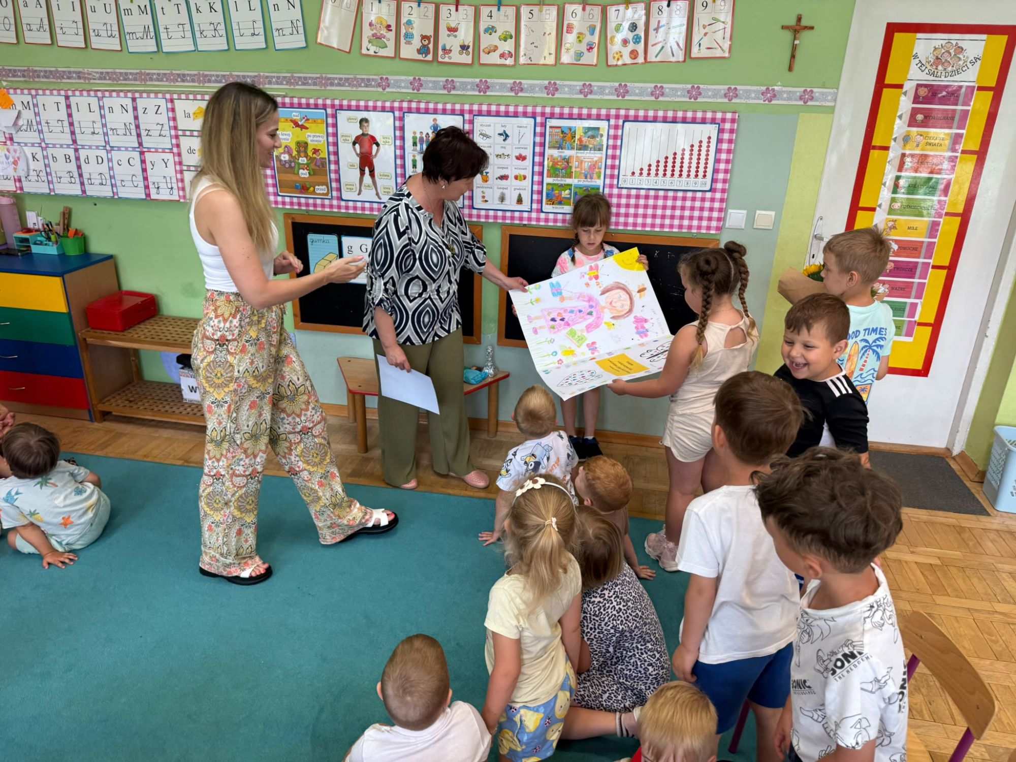Pani bibliotekarka wraz z dziećmi ogląda i czyta treść otrzymanej laurki.