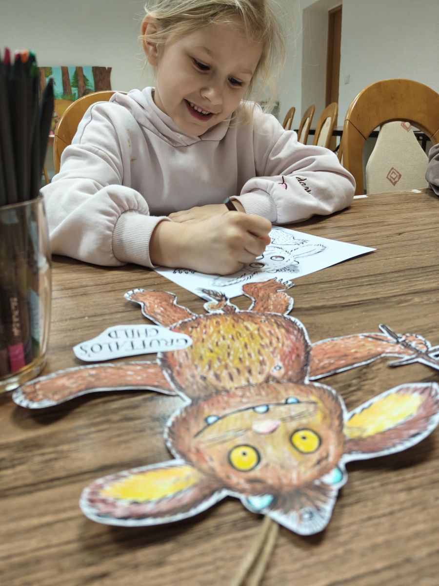 Uśmiechnięta dziewczynka kolorująca kolorowankę, przed nią leży na stole postać z papieru Dziecko Gruffalo
