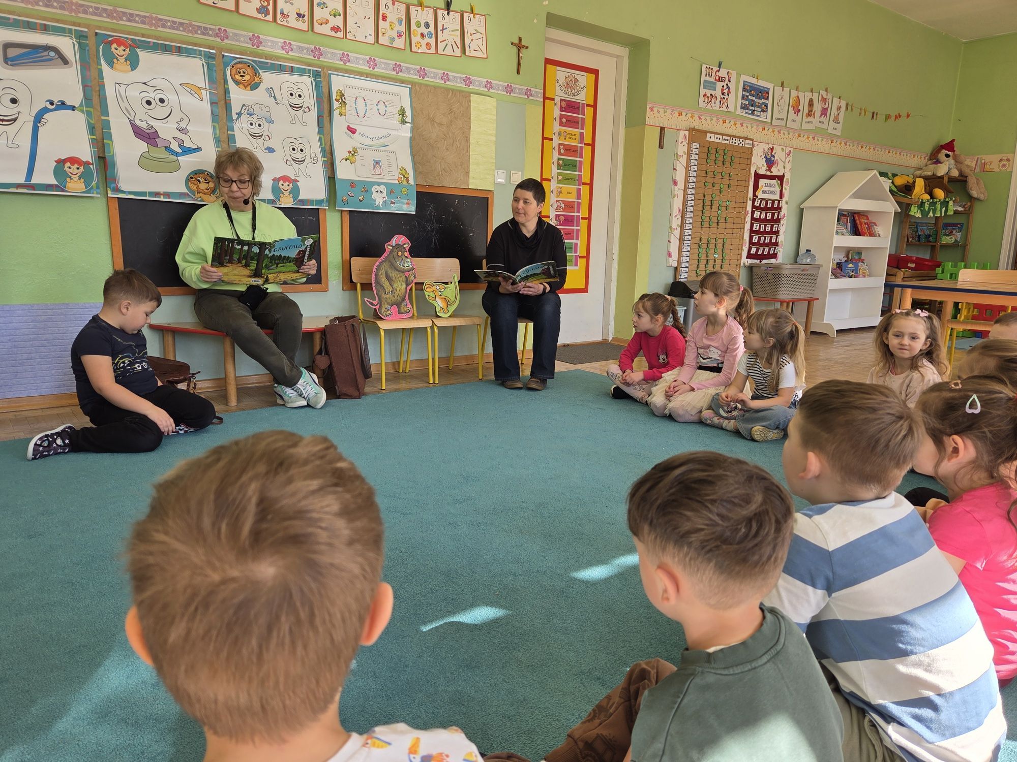 Dzieci siedzą w kole na dywanie i słuchają pań kt&oacute;re im czytają.