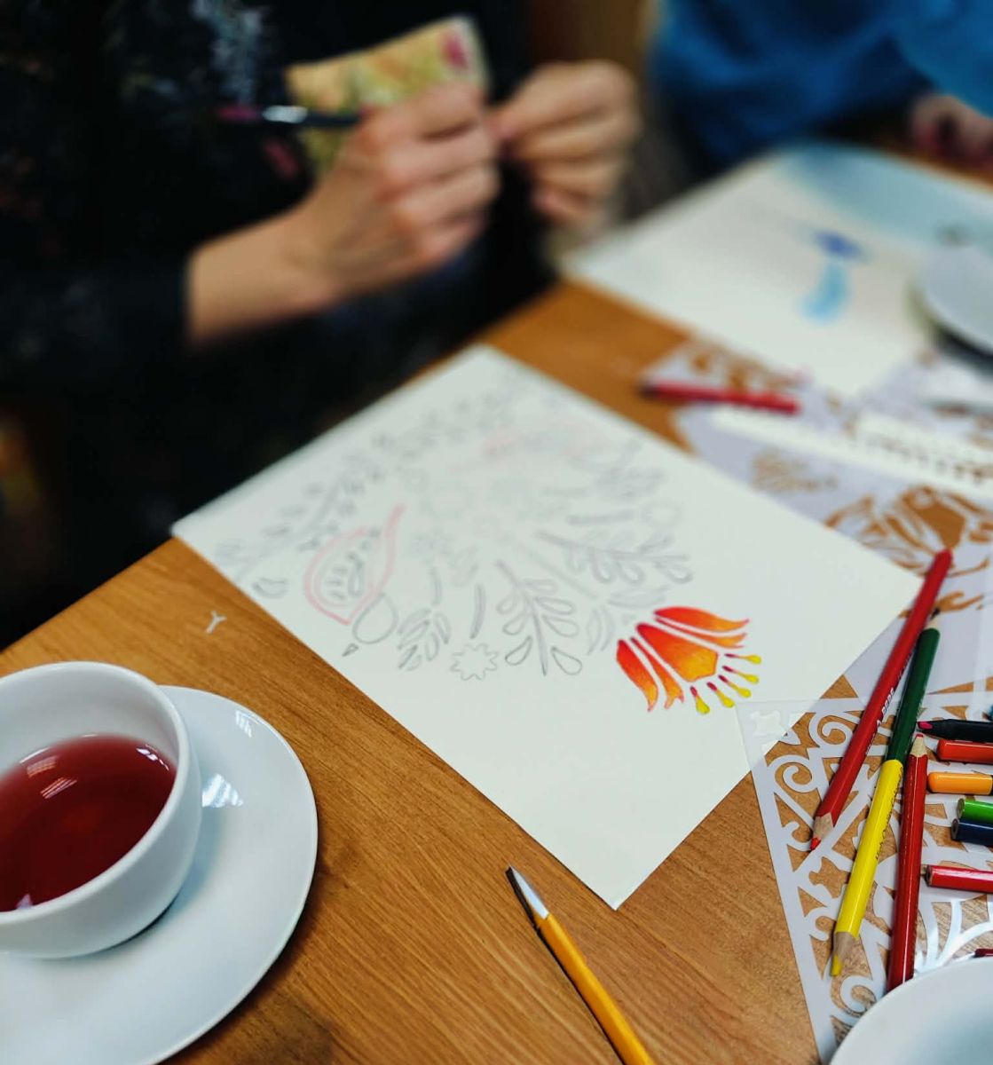 Na stole leży kartka papieru z rozpoczętą ilustracją ze wzorem w stylu folkowym. Widać ręce osoby rysującej.