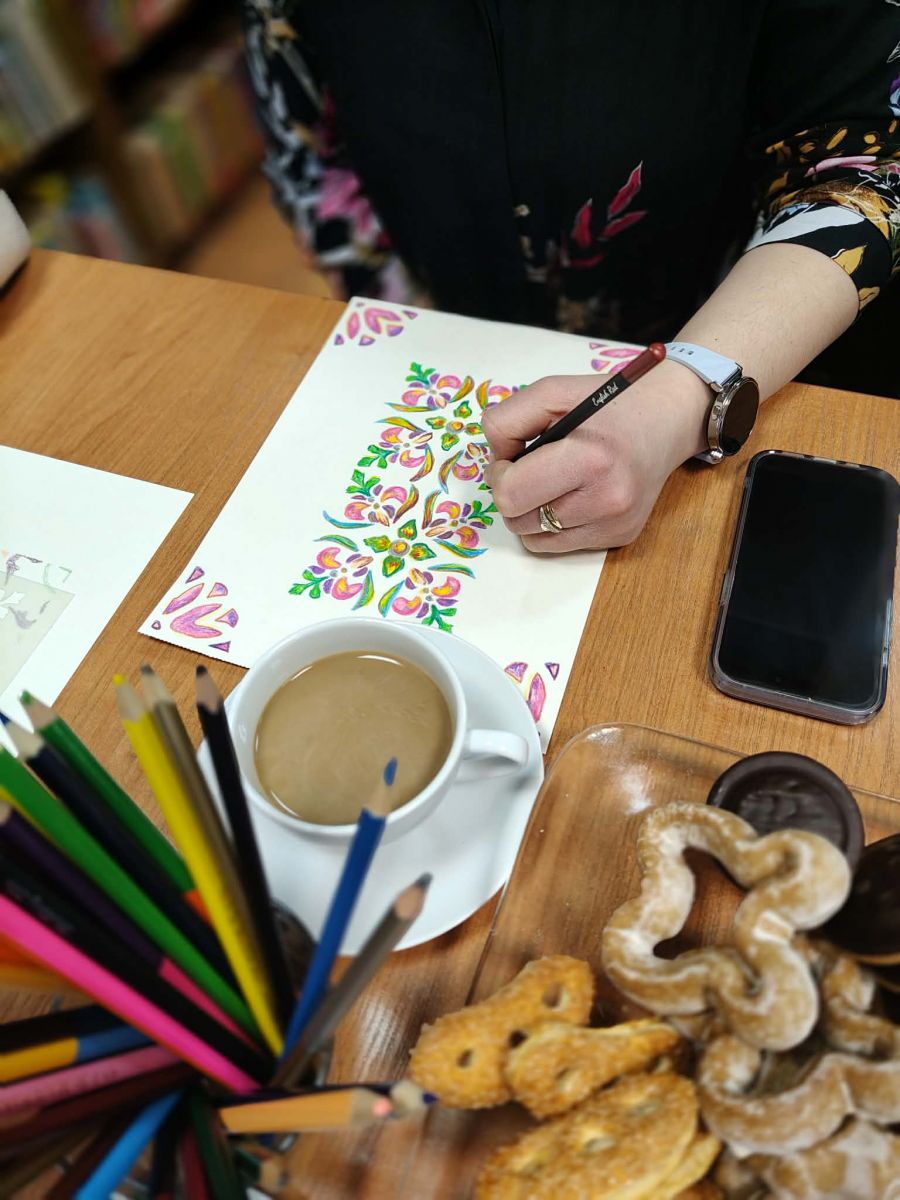 Na stole leży kartka papieru z rozpoczętą ilustracją ze wzorem w stylu folkowym. Widać ręce osoby rysującej, filiżankę z kawą, kredki i ciastka.