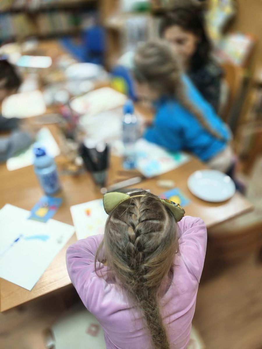 Dziewczynka widoczna tyłem siedzi przy stole na kt&oacute;rym leżą kartki papieru i przybory do rysowania.