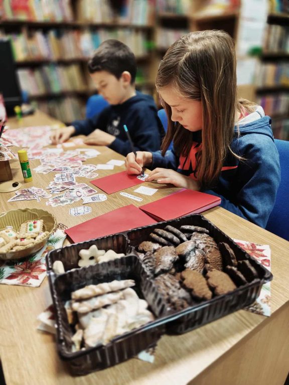Na zdjęciu widać dwoje dzieci siedzących przy stole w bibliotece i pracujących nad świątecznymi kartkami. Dziewczynka na pierwszym planie skupiona jest na rysowaniu lub ozdabianiu czerwonej kartki. Obok niej, nieco w tle, chłopiec r&oacute;wnież wykonuje własną kartkę, otoczony wyciętymi ozdobami i materiałami plastycznymi.