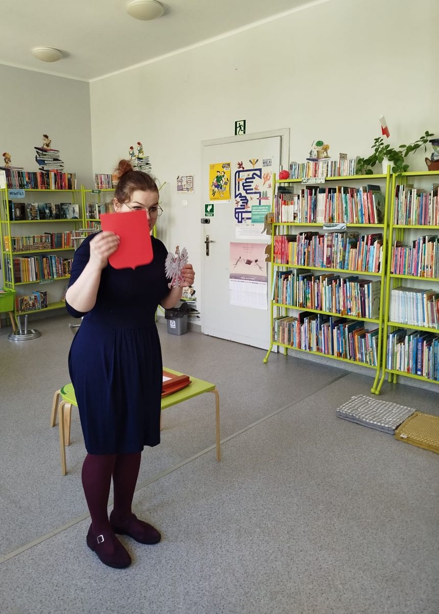 Bibliotekarka pokazuje dzieciom jak powstało godło Polski. Trzyma orła i osobno czerwony herb.