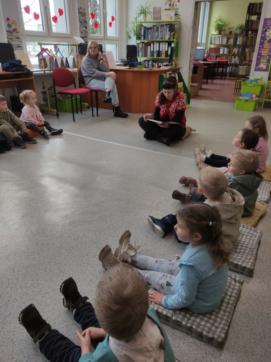 Pani czyta książkę zwr&oacute;cona do dzieci, kt&oacute;re siedża w k&oacute;łeczku i słuchają bajki o Bobusiu.
