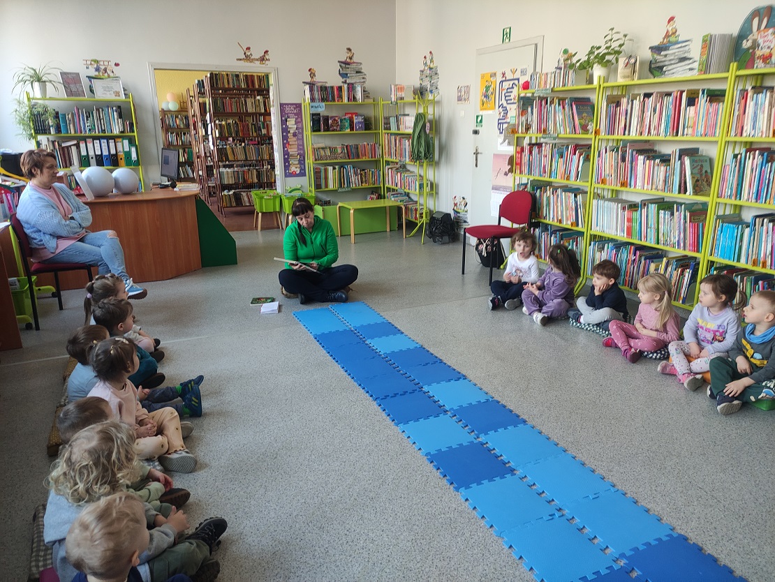 Pani czyta książkę pn. "Witaj żabko", zwr&oacute;cona twarzą do grupy przedszkolak&oacute;w.