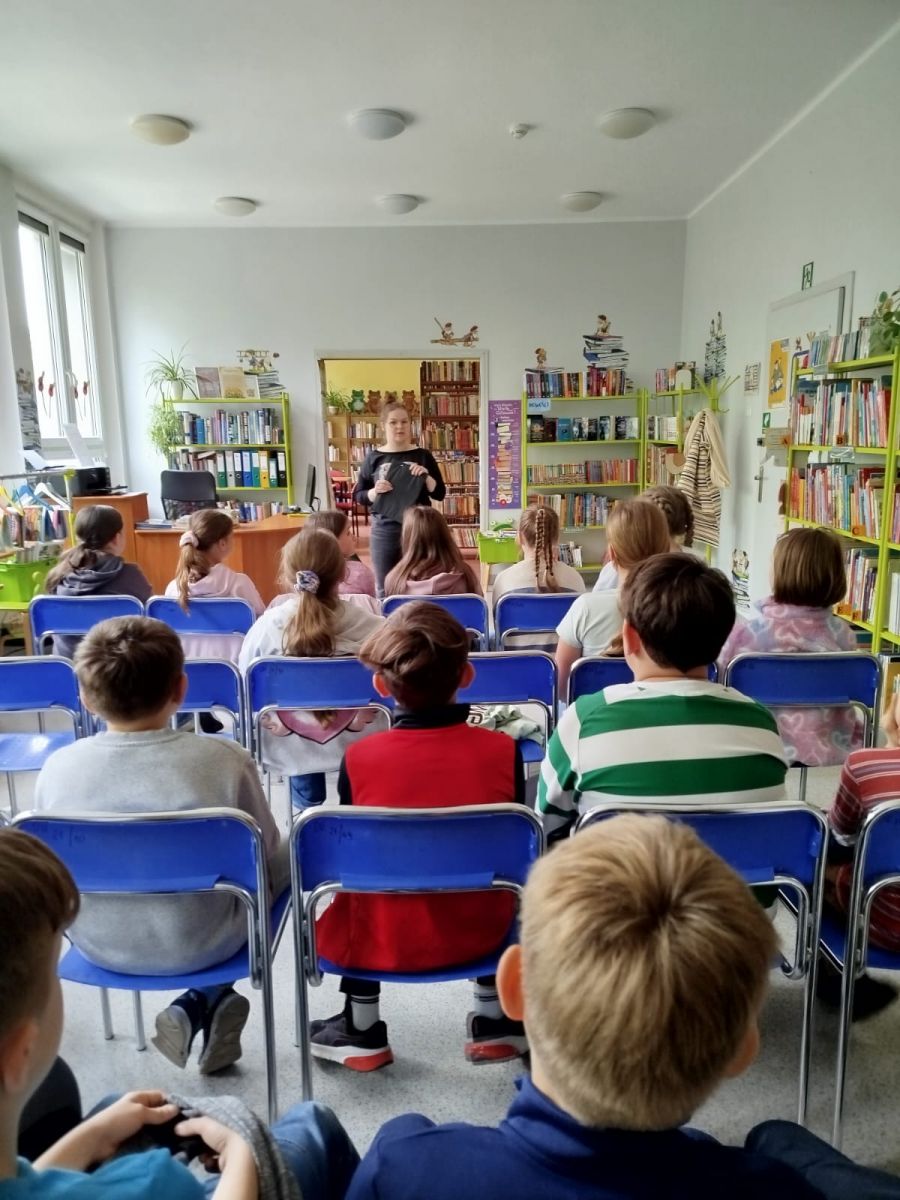 Bibliotekarka stoi przed grupa dzieci i omaiwa z nimi podstawowe zasady panuące w bibliotece.