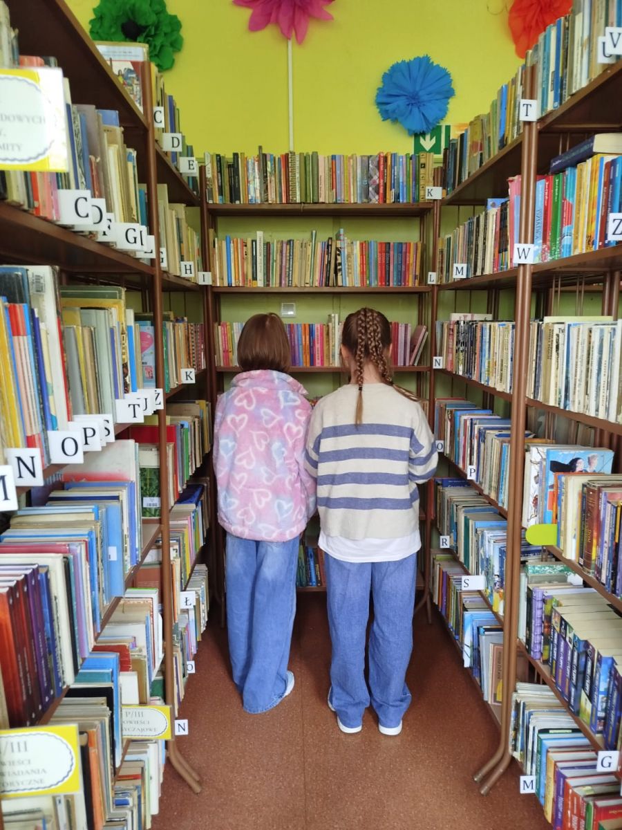 Dwie dziewczynki zapoznają się z tytułami dostępnymi na p&oacute;łkach w bibliotece.