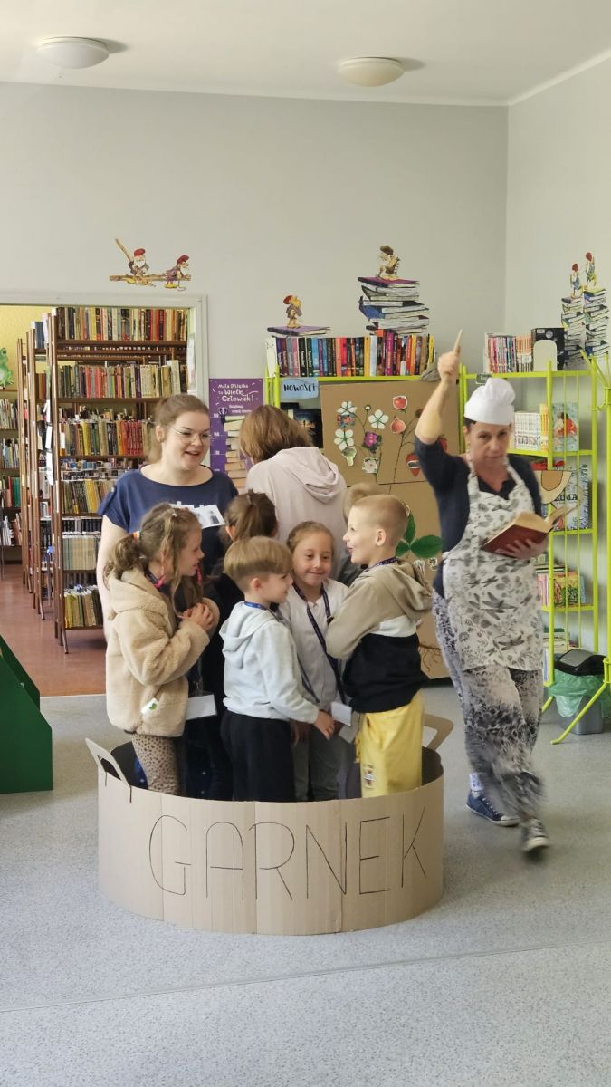 Bibliotekarka przebrana za kucharke biega wok&oacute;ł kartonowego garnka. W garnku stoi grupa przedszkolak&oacute;w.