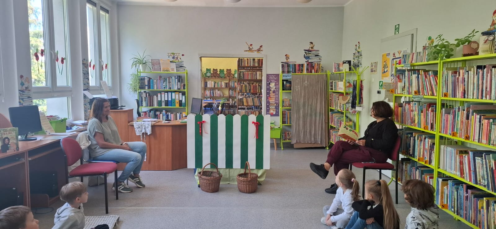 Na środku sali stoi, wykonany z kartonu stragan, nad nim widać kalarepkę. Obok straganu siedzi bibliotekarka i czyta wiersz.