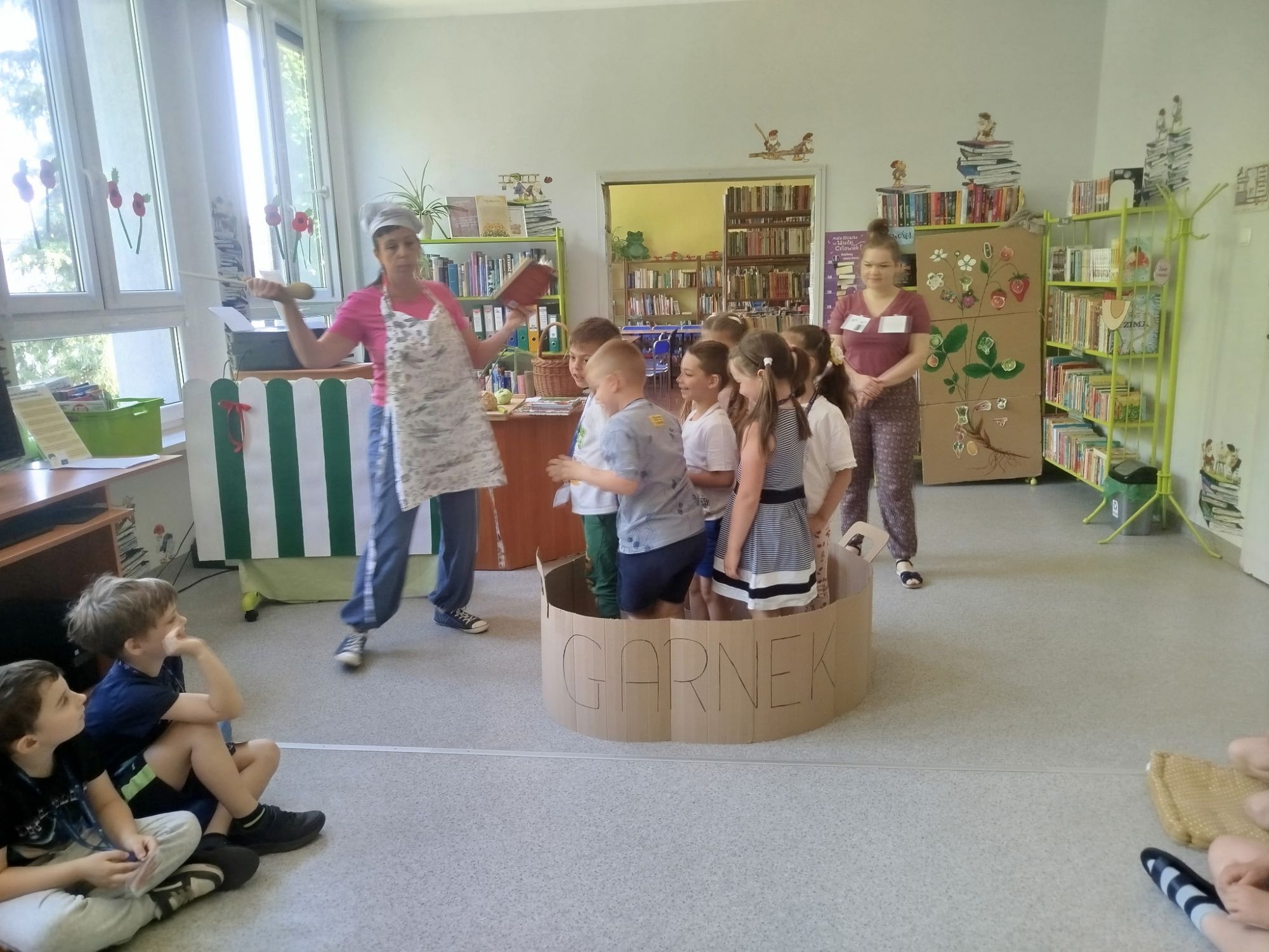 Bibliotekarka przebrana stoi z książką kucharską i drewnianą łyżką obo kartonowego garnka. W garnku stoi kilka przedszkolak&oacute;w.