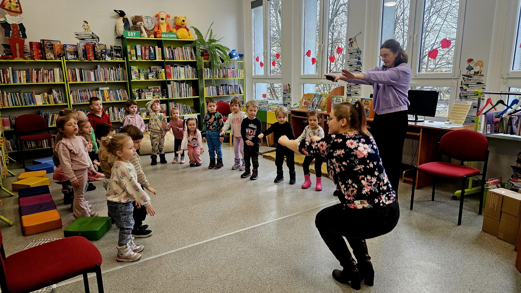 Przedszkolaki tańczą wraz z dwiema dorosłymi opiekunkami taniec robot&oacute;w pokazywany przez bibliotekarkę