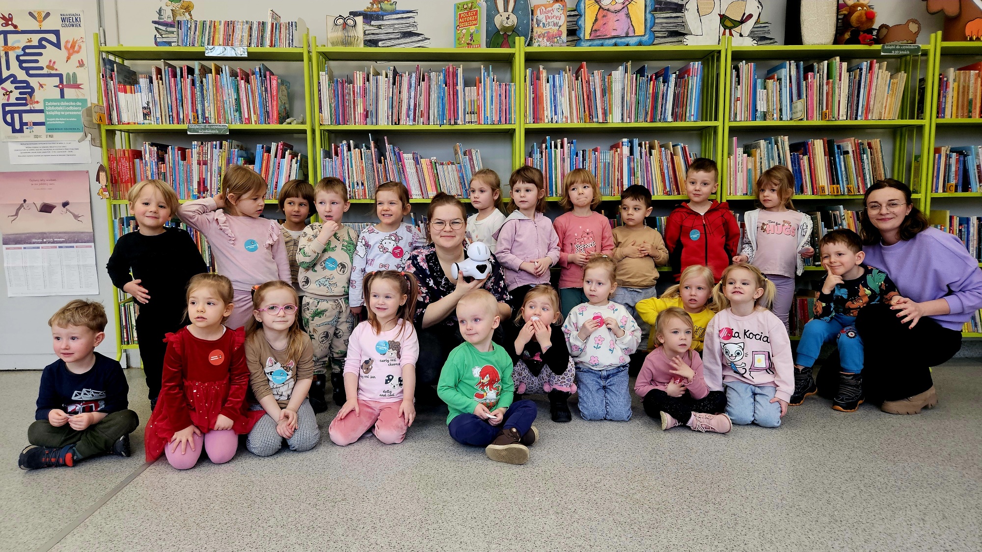 Grupa przedszkolak&oacute;w na tle regał&oacute;w z książkami z bibliotekarką i robotem Photonem.