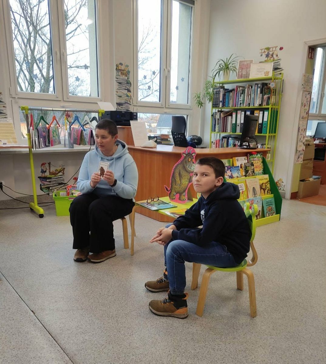  Dwie osoby siedzą przy małym stoliku, jedna osoba pokazuję kartę a druga słucha.