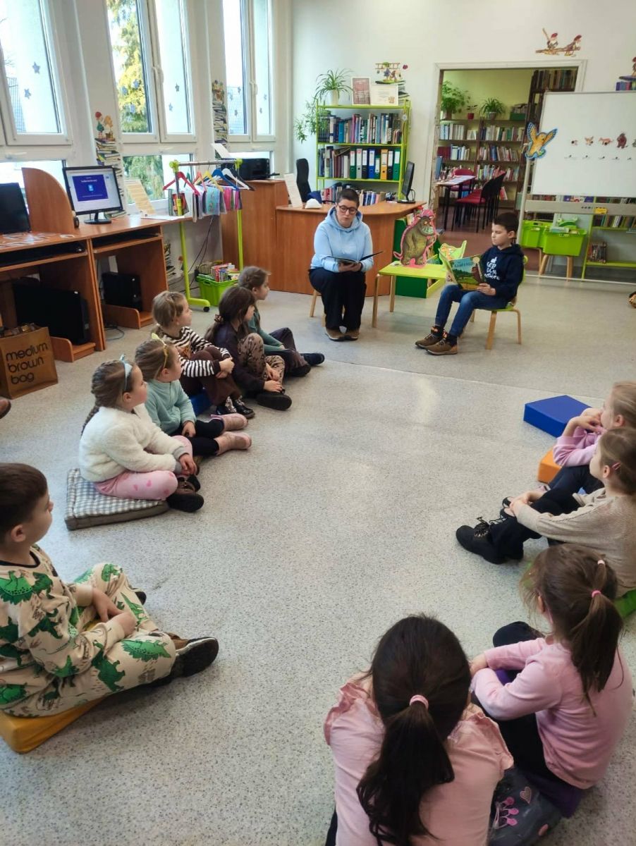 Dwie osoby siedzą przy małym stoliku i czytają książki grupie dzieci siedzącej przed nimi na podłodze.